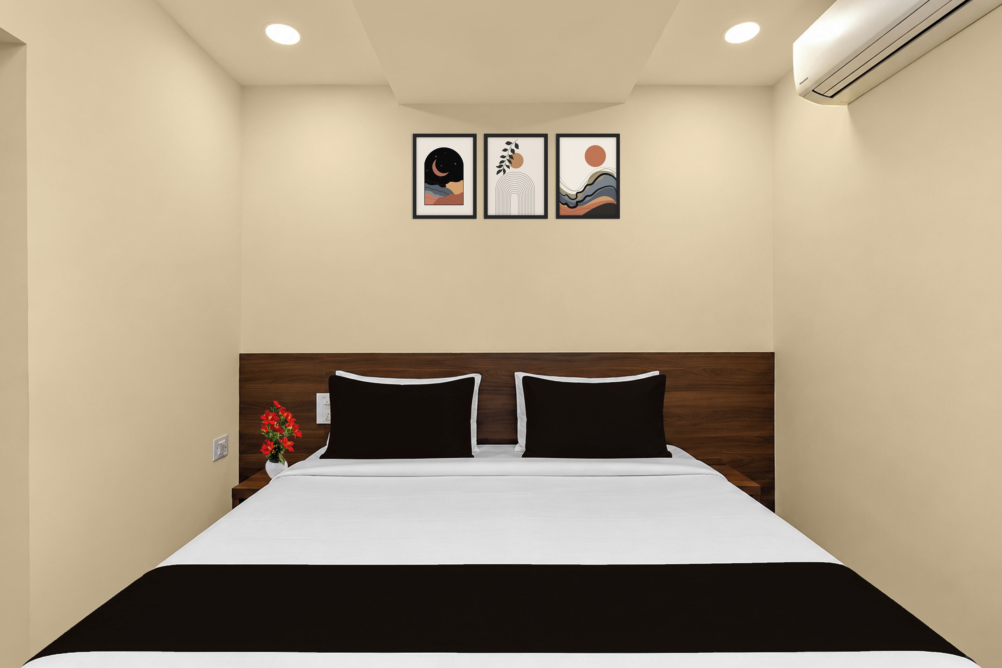 A hotel room with a bed, a nightstand, a vase with flowers, and three framed artworks on the wall.