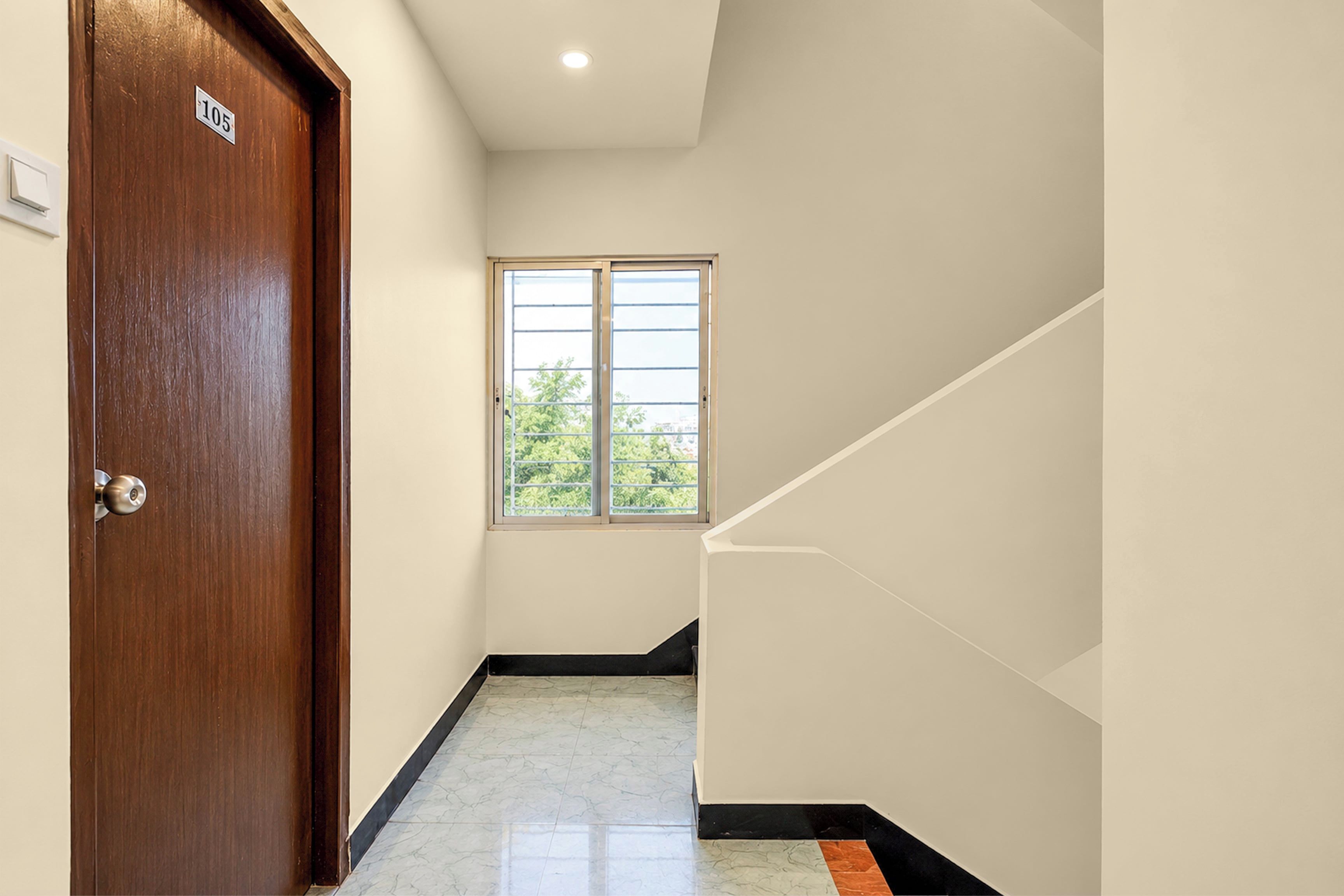 Un pasillo con una puerta de madera marcada '105', una ventana con vista a zonas verdes y un suelo de baldosas con borde negro.