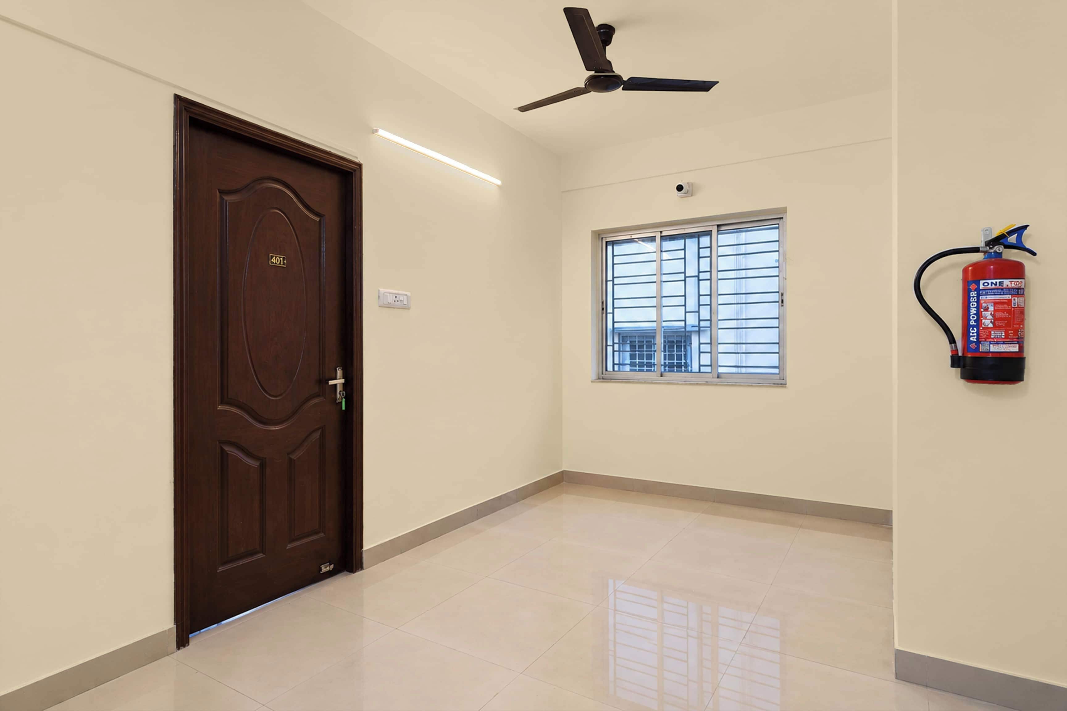 A hallway with a brown door, a ceiling fan, a window with bars, and a fire extinguisher mounted on the wall.