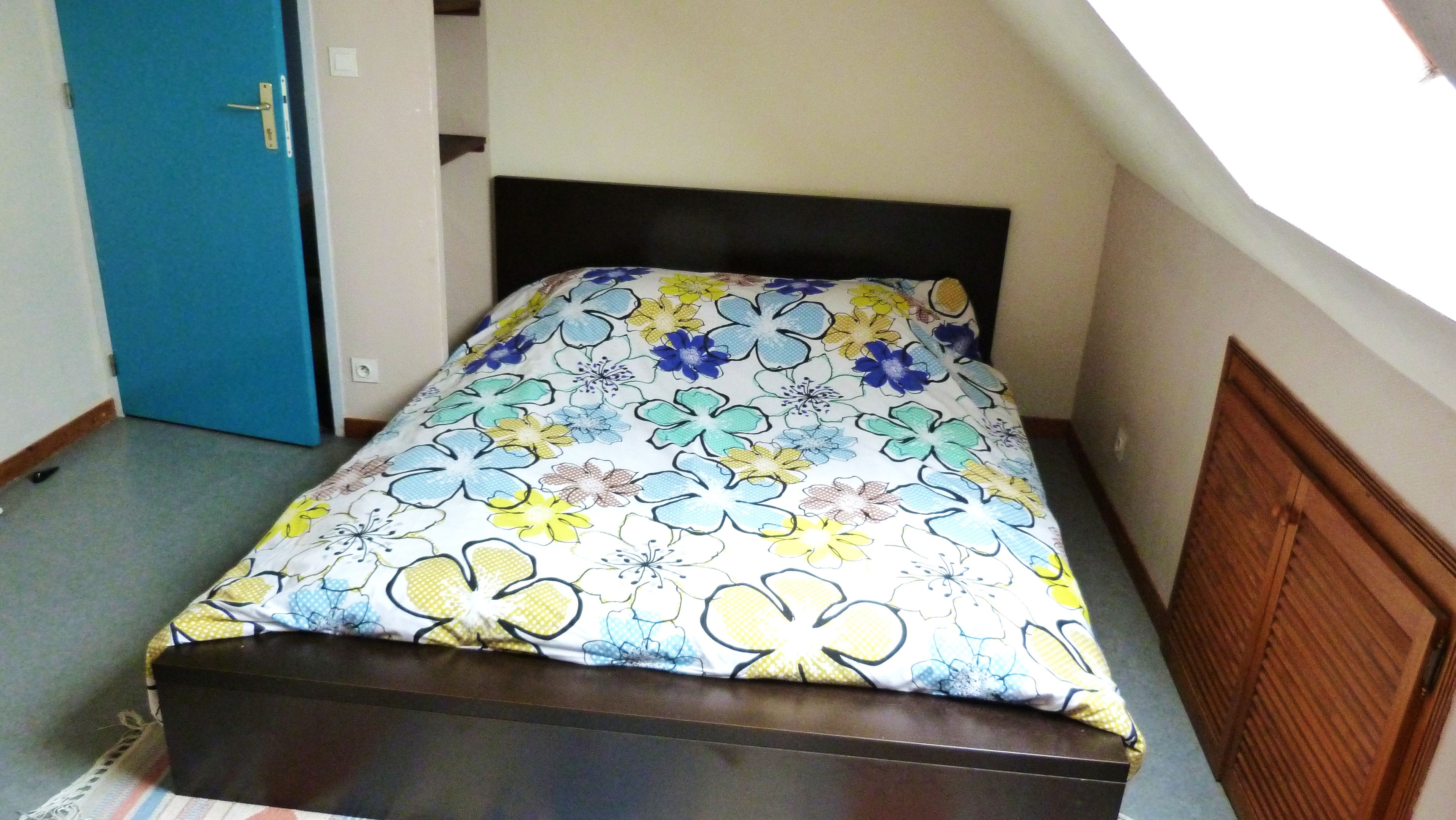 Un lit simple avec une couvre-lit à motifs floraux, dans une pièce dont la porte est bleue et qui possède une tête de lit en bois.