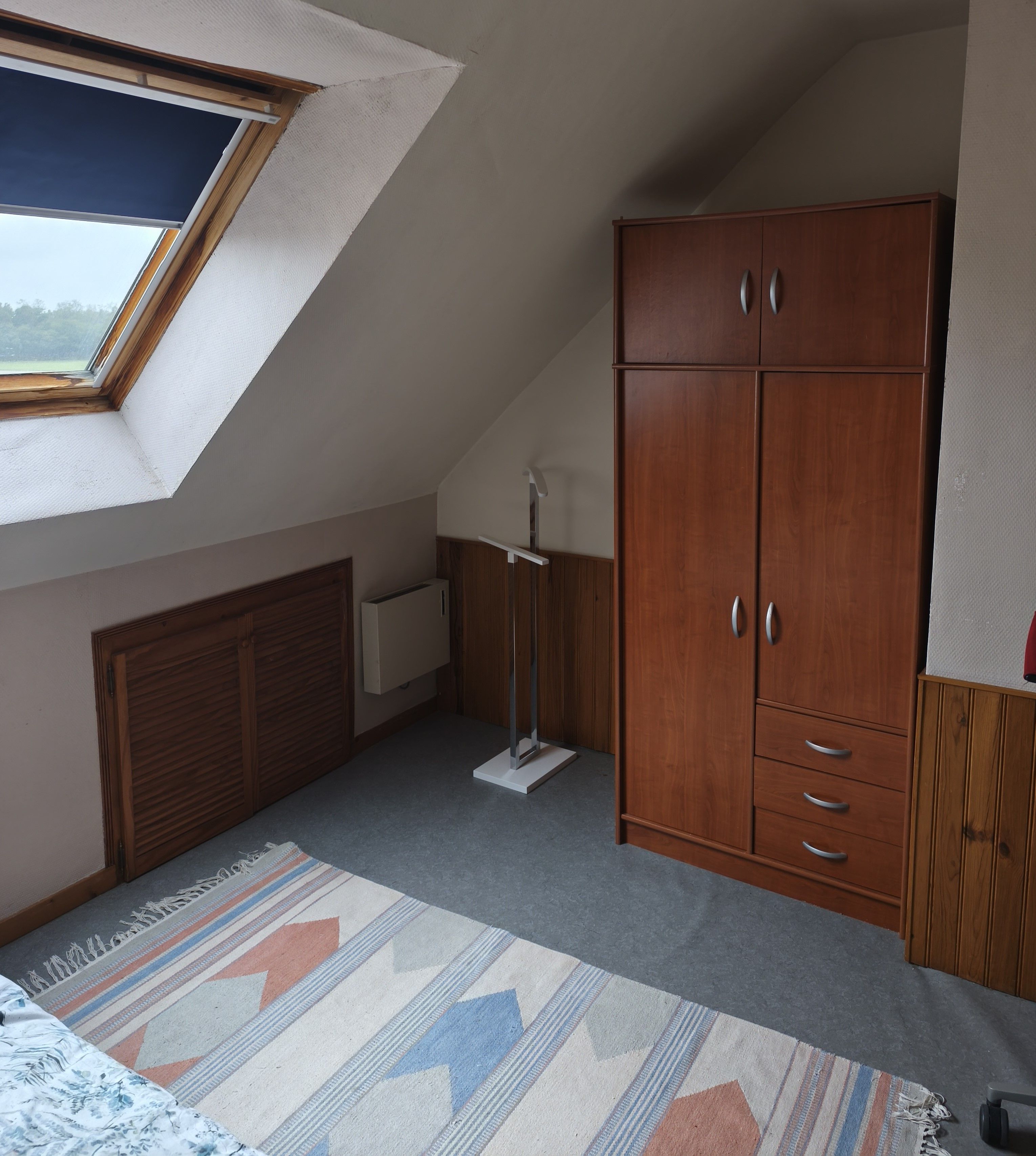 Une chambre avec un plafond en pente, une armoire, un radiateur et une fenêtre.