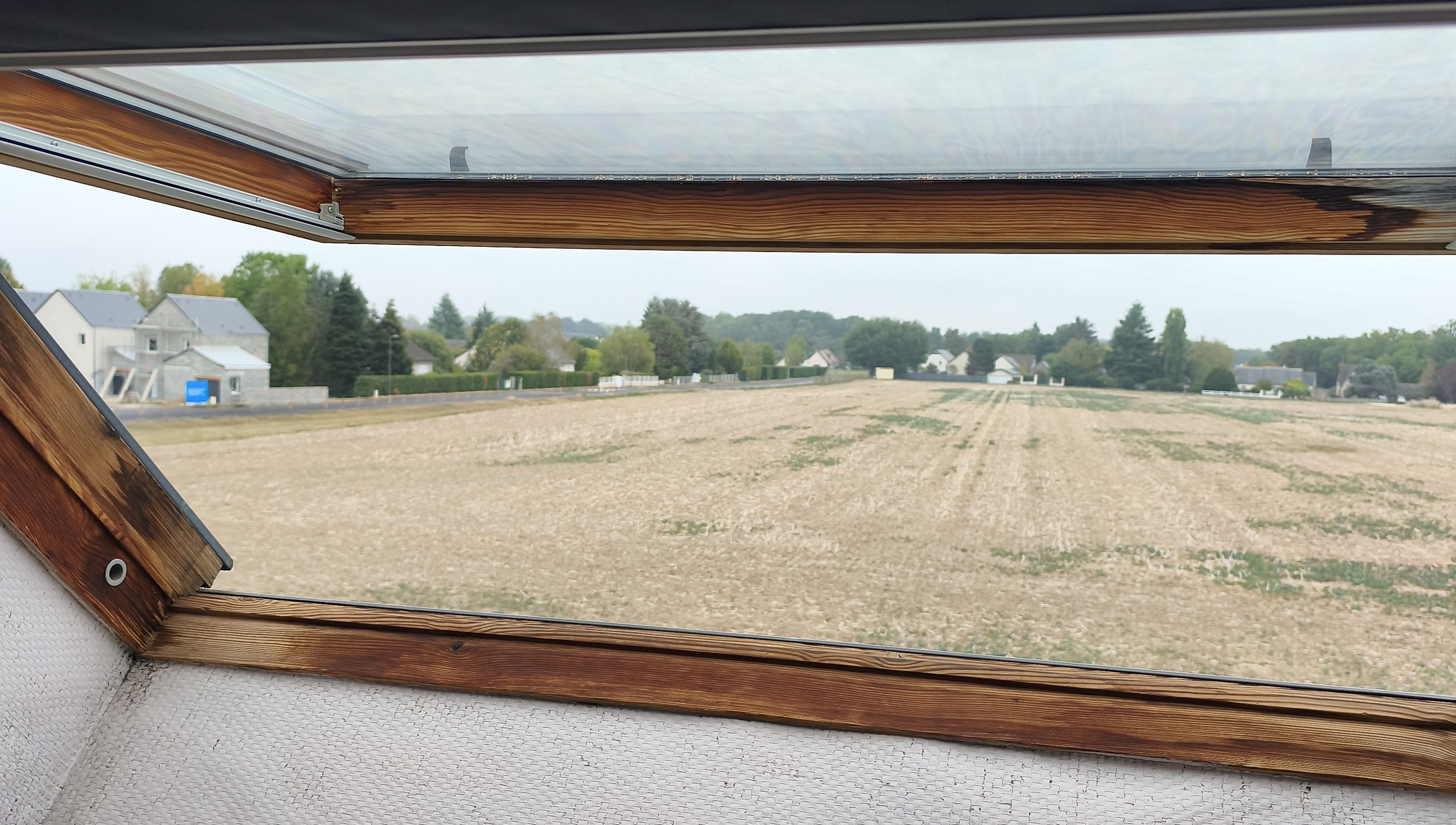 Une vue à travers une fenêtre à cadre en bois, donnant sur un champ avec des maisons en arrière-plan.