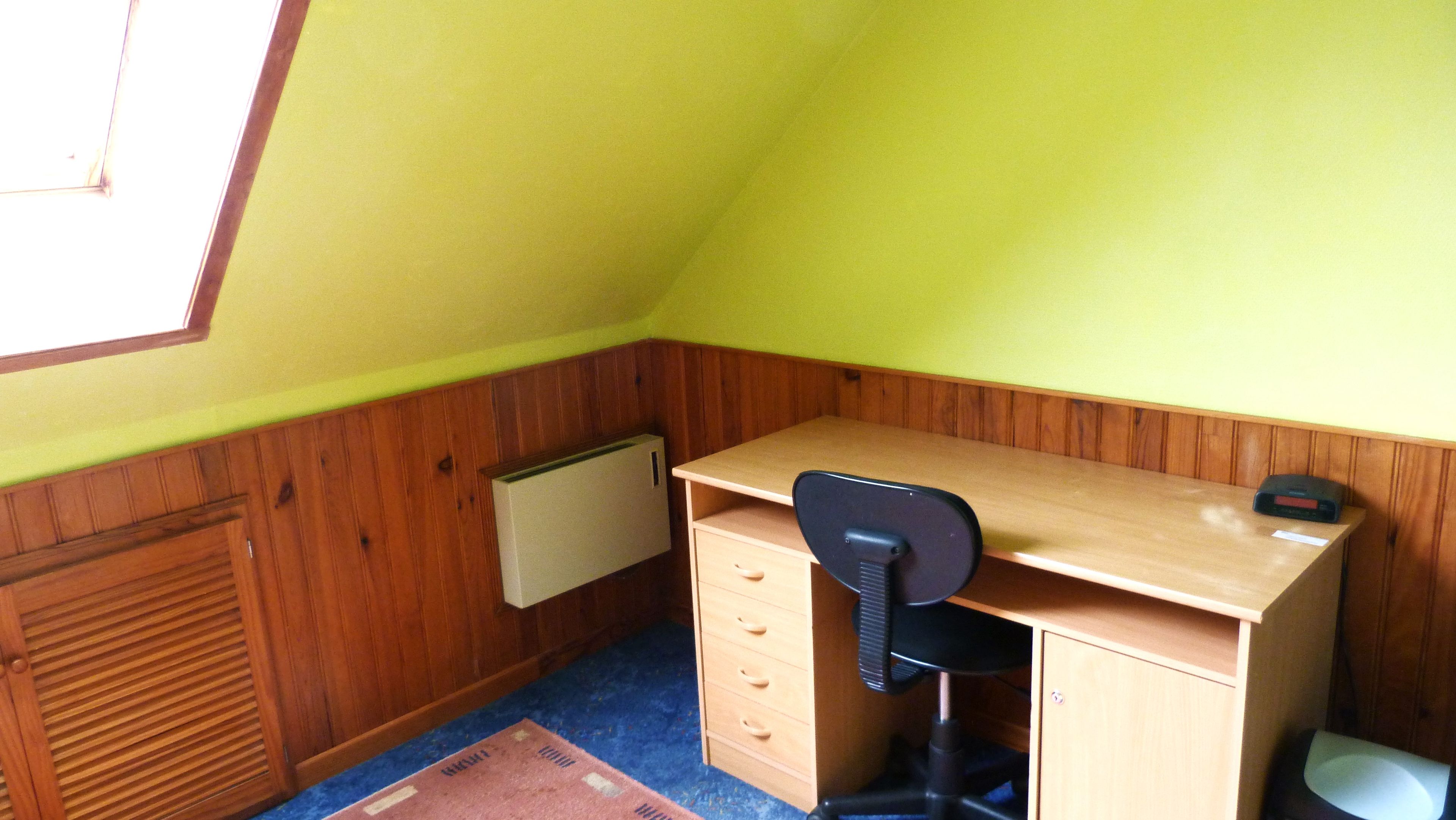 A small room with a sloped ceiling, a wooden desk with a chair, a radiator, and a window.