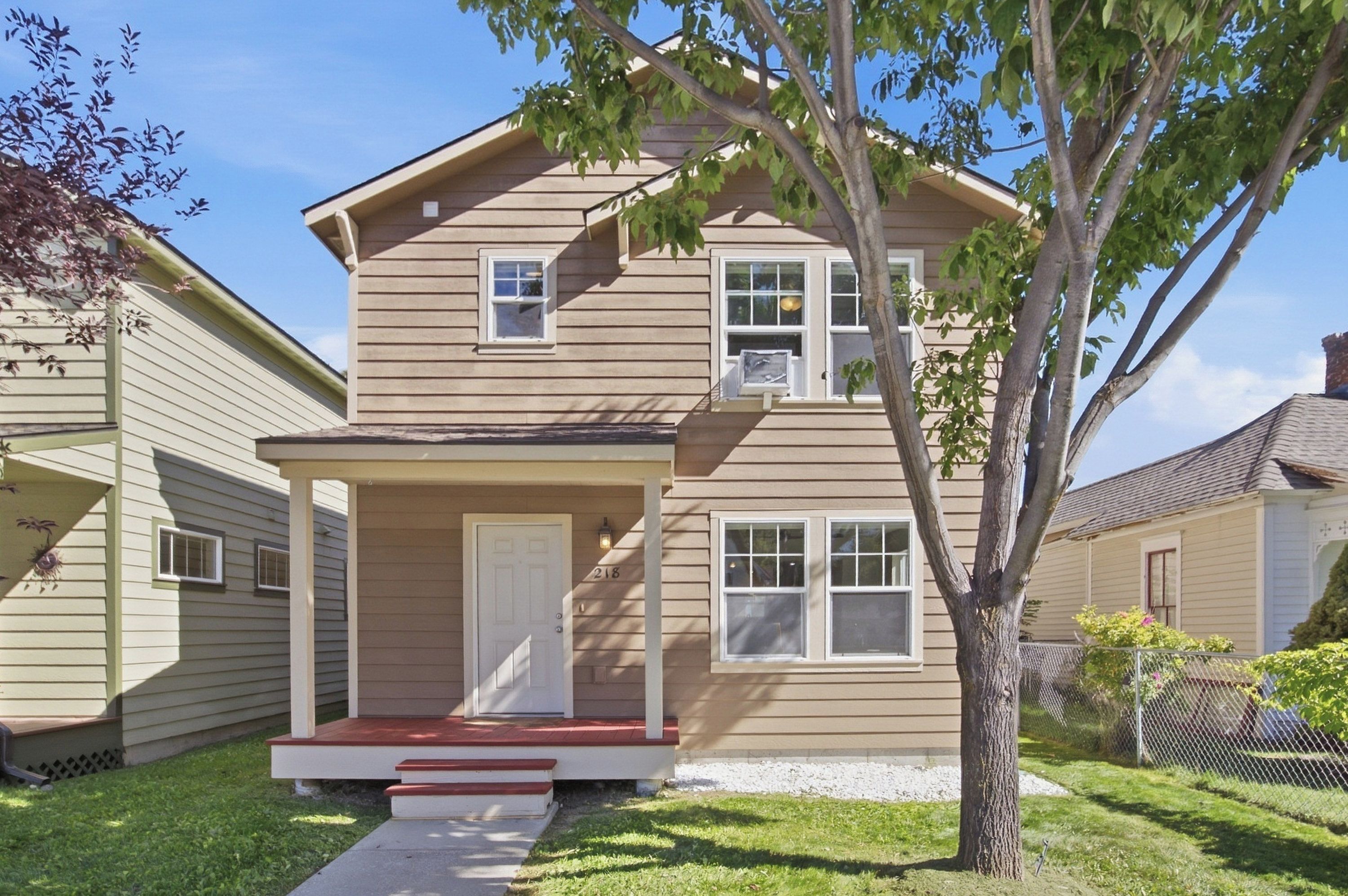 A two-story residential house with a front porch, a small attached garage, and a tree in the front yard.