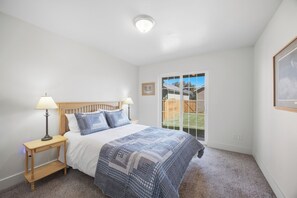 A bedroom with a bed, two bedside lamps, a wooden nightstand, a carpeted floor, a ceiling light, a window with a view of a backyard, and a framed picture on the wall.