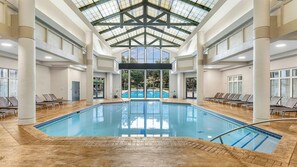 Une piscine intérieure dotée d’un toit en verre, entourée de chaises longues et offrant une vue sur une piscine extérieure.