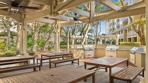 Une terrasse extérieure avec des tables et des bancs en bois, un ventilateur de plafond et un espace barbecue.