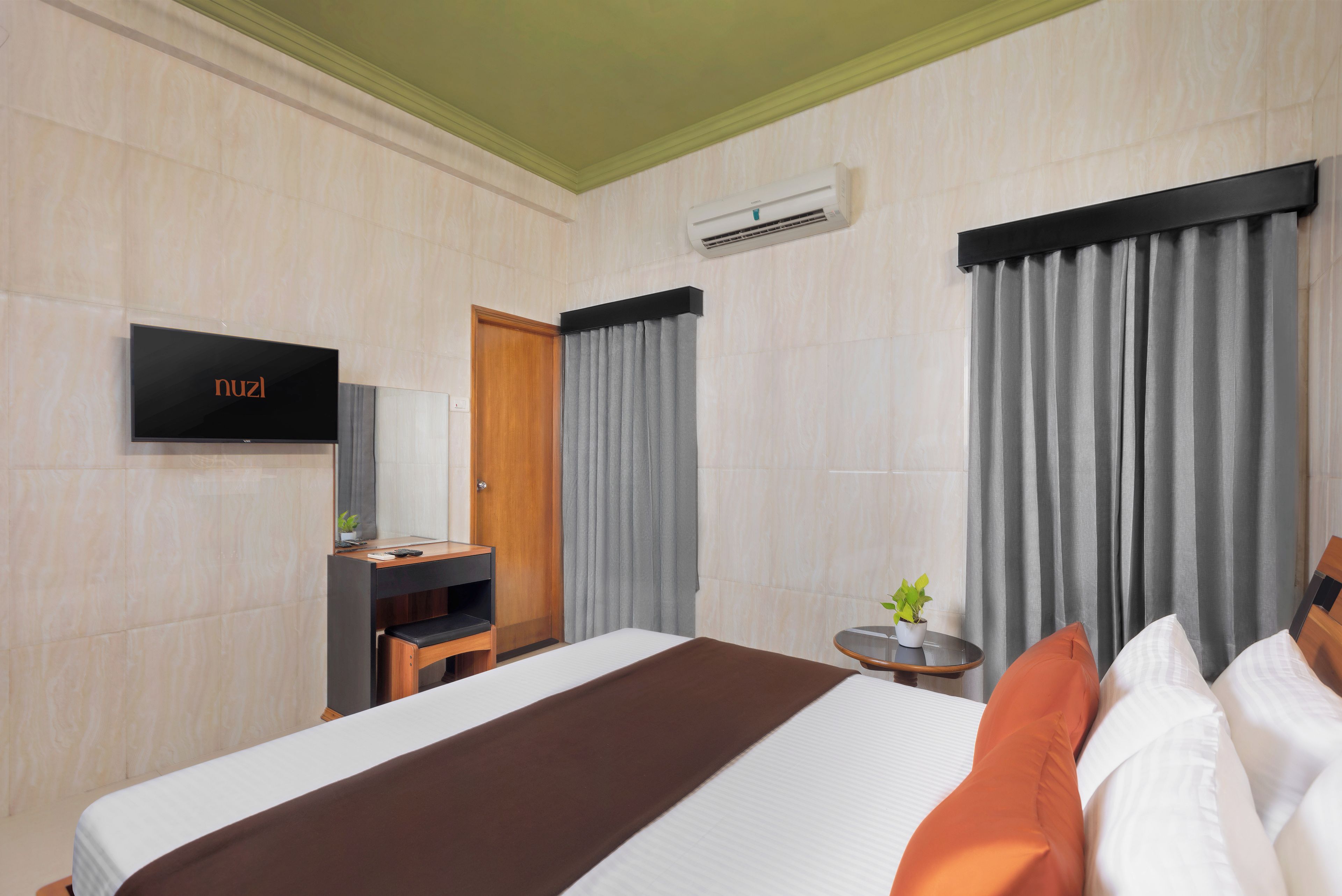 A hotel room with a bed, a television, a desk, a chair, a small table with a plant, and curtains.