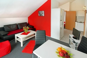 A modern living room with a red accent wall, a black leather sofa, a white coffee table, and a dining area with a fruit bowl. - 2-room apartment with a fantastic view (Winterberg)