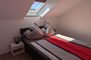 Quarto com claraboia, cama com roupa de cama listrada e mesa de cabeceira com abajur.