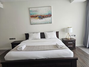 A neatly made bed with white linens and a folded towel, a bedside table with a lamp, and a painting on the wall. - Cozy 1-bedroom apartment in serene Nairobi with fitness room (Nairobi)