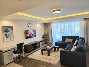 A modern living room with a sofa, TV, desk, and a large window with curtains. - Cozy 1-bedroom apartment in serene Nairobi with fitness room (Nairobi)
