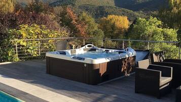 Un bain à remous sur une terrasse en bois, avec une piscine, le tout entouré d’arbres et de collines.