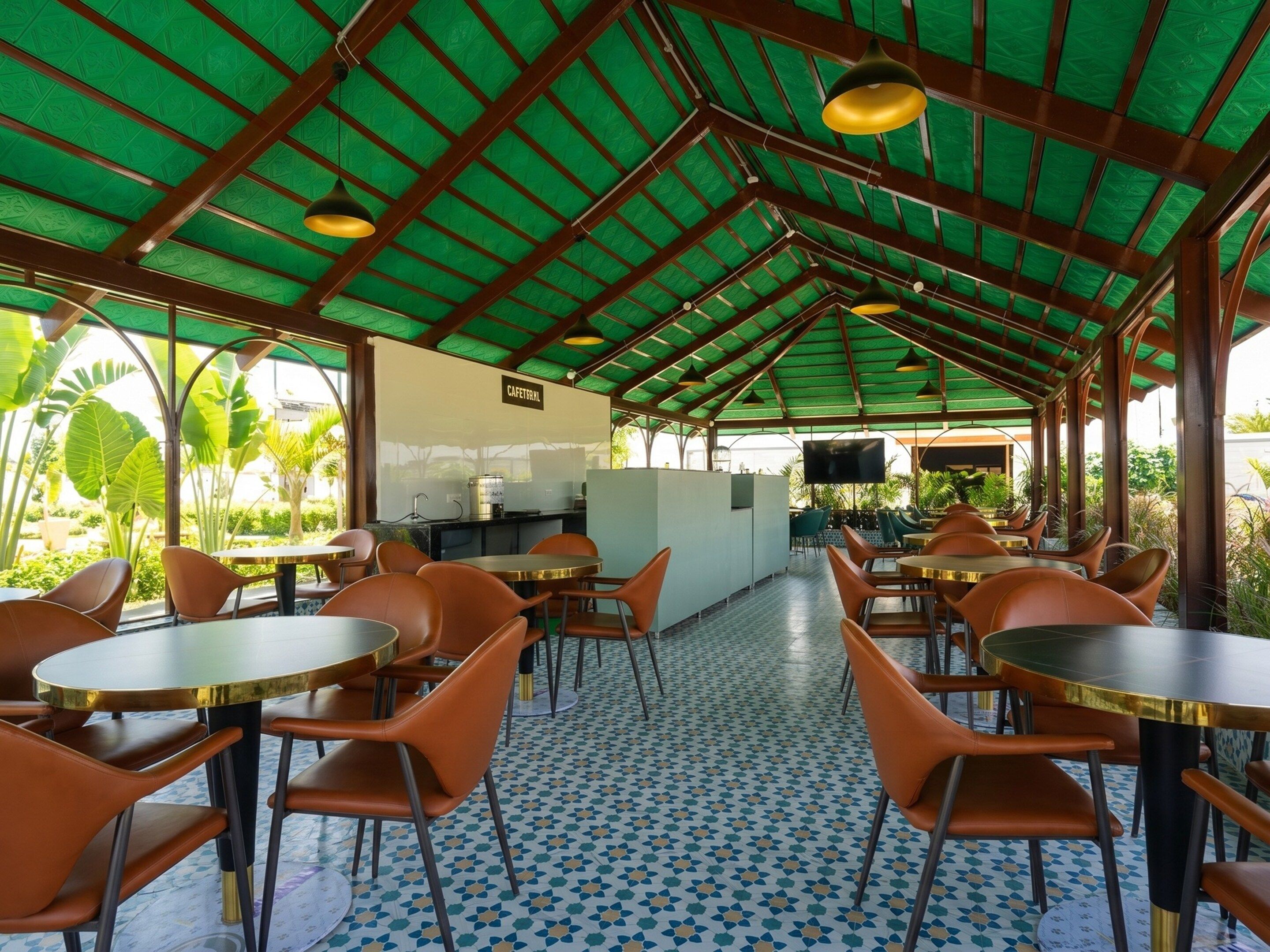 A covered outdoor seating area with tables and chairs, a kitchen area, and a view of greenery.