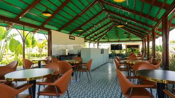 A covered outdoor seating area with tables and chairs, a kitchen area, and a view of greenery.