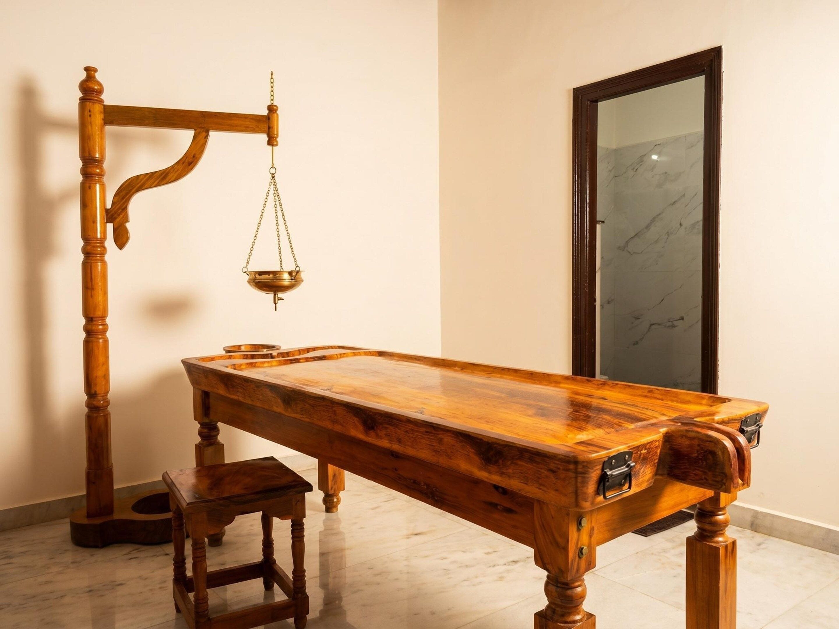 A wooden massage table with a footrest and a wooden chair, a hanging brass scale, and a marble-tiled floor.