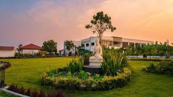 A serene garden with a Buddha statue, lush greenery, and a modern building in the background.