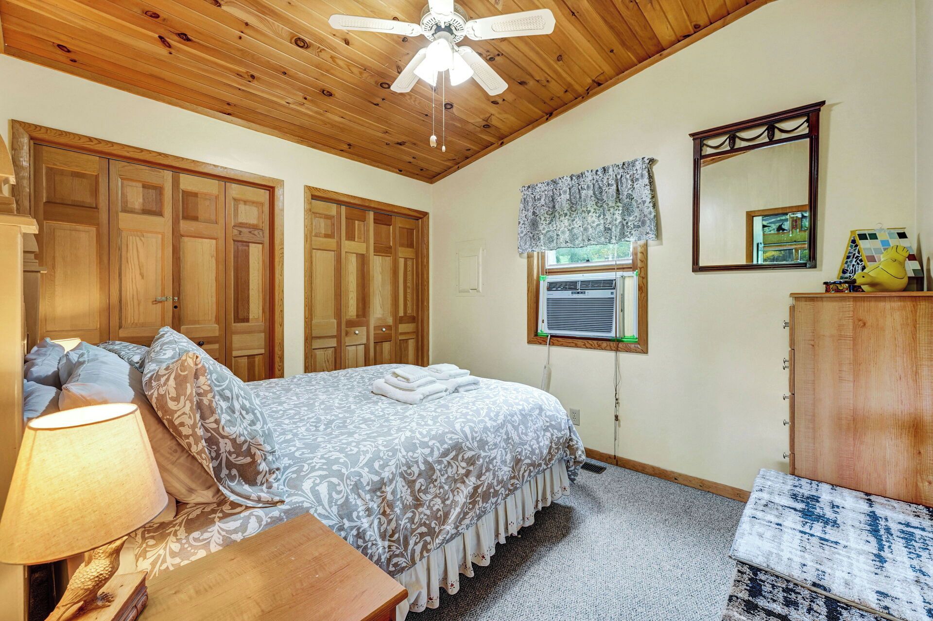 Une chambre à coucher comprenant un lit, une table de chevet, une lampe, un ventilateur de plafond, des armoires en bois, une fenêtre avec un climatiseur, un miroir et une commode.
