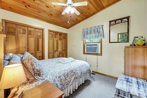 A bedroom with a bed, a nightstand, a lamp, a ceiling fan, wooden wardrobes, a window with an air conditioner, a mirror, and a dresser.