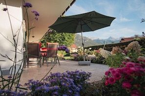 Ein Sitzbereich im Freien mit Tisch und Stühlen unter einem großen Sonnenschirm, umgeben von verschiedenen Blumen und Blick auf Berge.