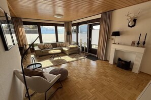 A spacious living room with a wooden floor, a sofa set, a fireplace, and a large window overlooking a scenic view. - Holiday home in Meschede with swimming pool (Meschede)