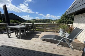 Um deck externo com mesa e cadeiras, uma poltrona de descanso e vista para uma paisagem montanhosa.