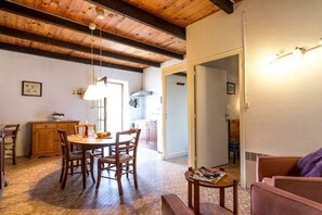 A dining area with wooden chairs and a table, a kitchen in the background, and a living area with a sofa and a small table. - Gite Louise - Mas Paraloup (Lagorce)