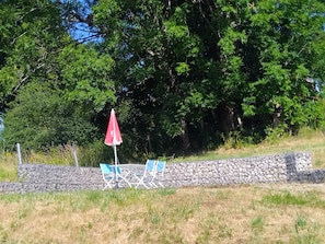 A stone wall with a table and chairs, an umbrella, and a grassy area with trees in the background. - La Tour Au Comté Vacation Rental. Experience the authentic lifestyle, from a 15th-century tower to farm life. (Charency)