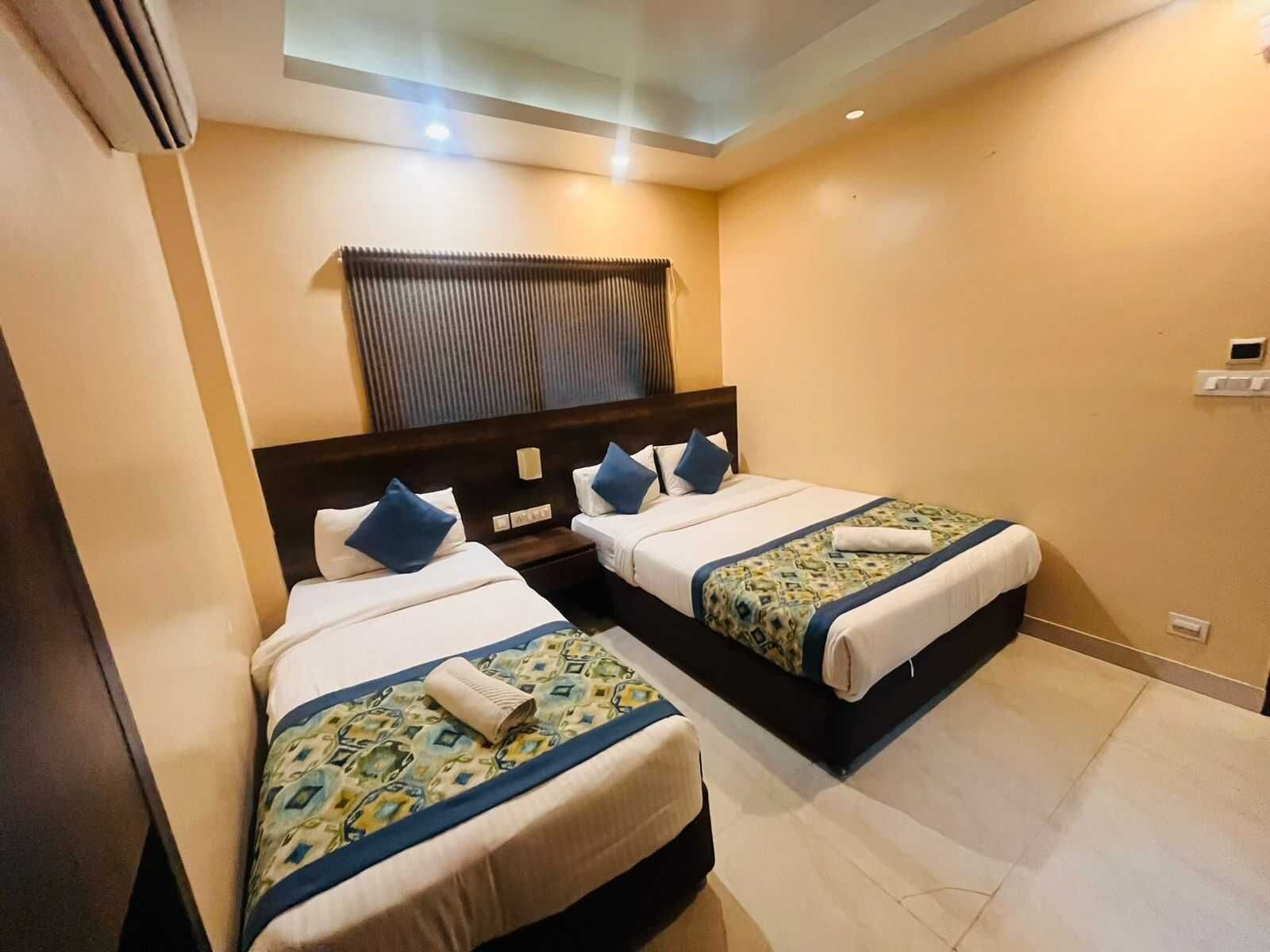 A hotel room with two beds, a window with blinds, a ceiling light, and a wall-mounted thermostat.