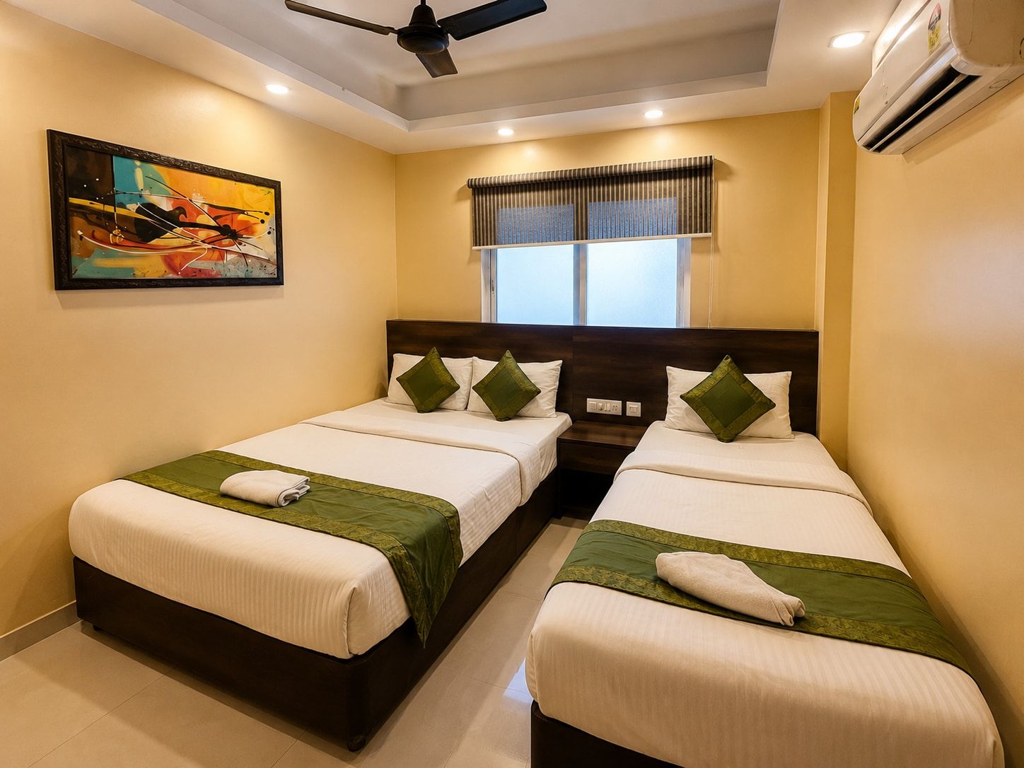 A hotel room with two beds, a ceiling fan, an air conditioning unit, a window with curtains, and a painting on the wall.