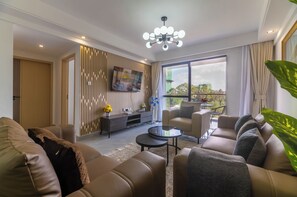 Ein modernes Wohnzimmer mit einer Sofagarnitur, einem Couchtisch, einem an der Wand montierten Fernseher und einem großen Fenster mit Blick ins Freie.