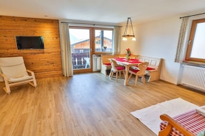 Een kamer met een houten wand, een eettafel met rode stoelen, een witte bank en een aan de muur bevestigde televisie.