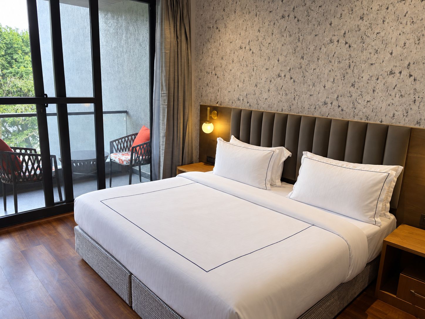 Una habitación de hotel con una cama grande, una mesita de noche, una lámpara de pared, un ventanal con cortinas y un balcón con una silla.