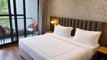 Una habitación de hotel con una cama grande, una mesita de noche, una lámpara de pared, un ventanal con cortinas y un balcón con una silla.