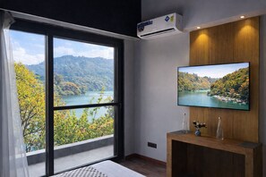Habitación con un amplio ventanal que ofrece una vista panorámica de un lago y un bosque, un televisor montado en la pared y un mueble consola de madera con objetos decorativos.