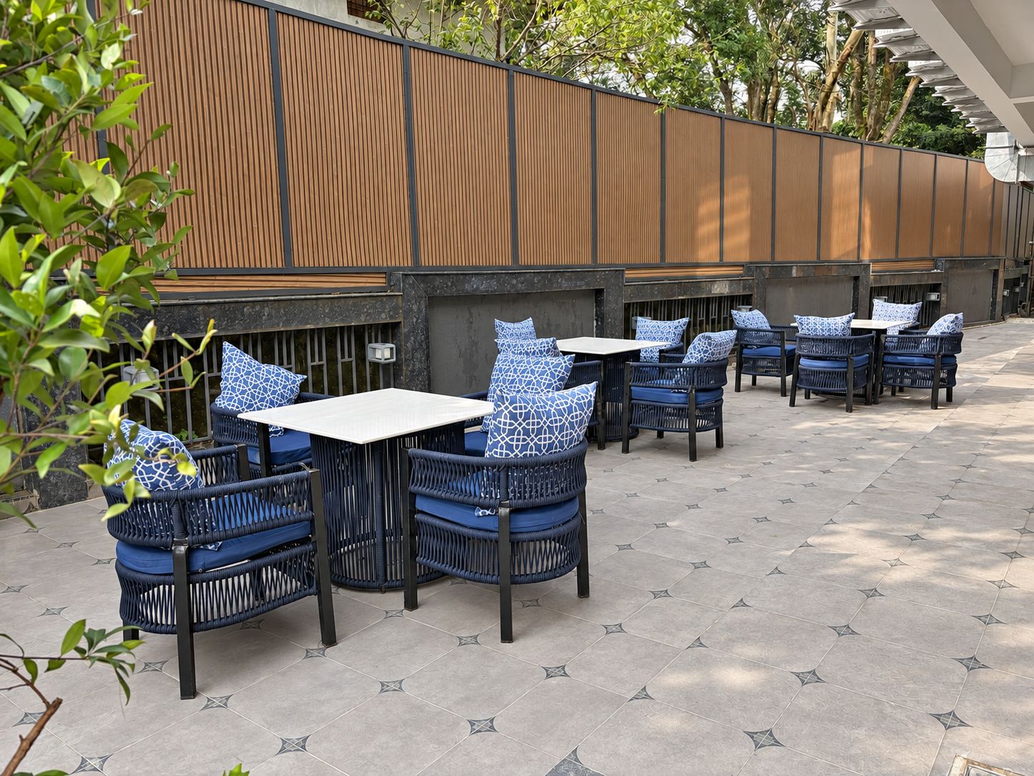 An outdoor seating area with wicker chairs and tables, a wooden fence, and a tiled floor.