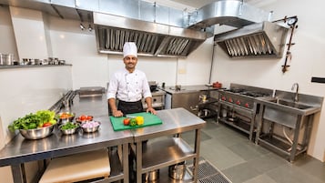 Un chef dans une cuisine professionnelle équipée d’appareils en acier inoxydable, avec une grande planche à découper recouverte de légumes et une variété d’ustensiles de cuisine.