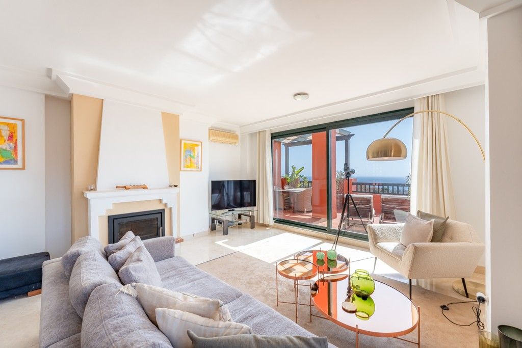 A modern living room with a sofa, television, and a fireplace. There is a balcony with a view of the sea.