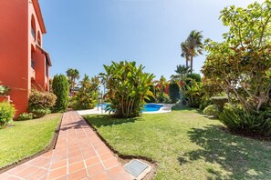 A pathway leading to a swimming pool surrounded by lush greenery and a red building. - Bonita Hills – Penthouse in Calahonda with Panoramic Sea Views & Large Terrace (Mijas)