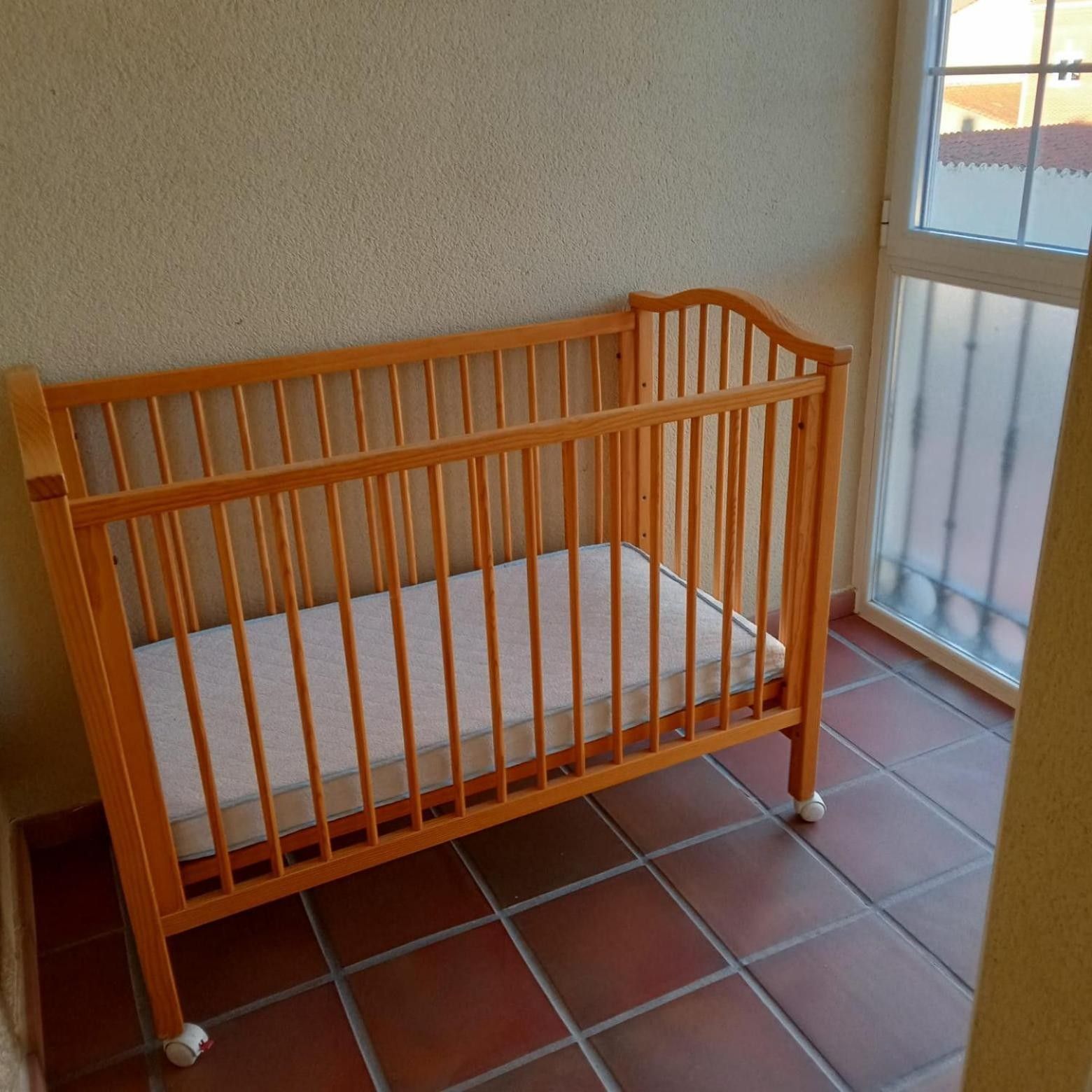 A wooden crib with a white mattress on a tiled floor, positioned near a window with a view of a building outside.