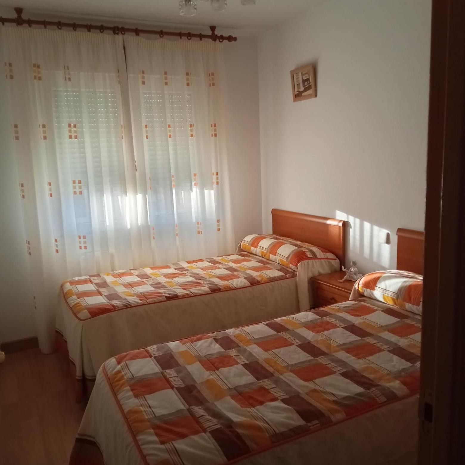 A room with two beds, a window with sheer curtains, and a small bedside table with a lamp.