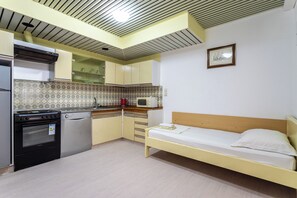 Cozinha compacta com geladeira, fogão e micro-ondas. Há uma cama com cabeceira de madeira e um travesseiro.