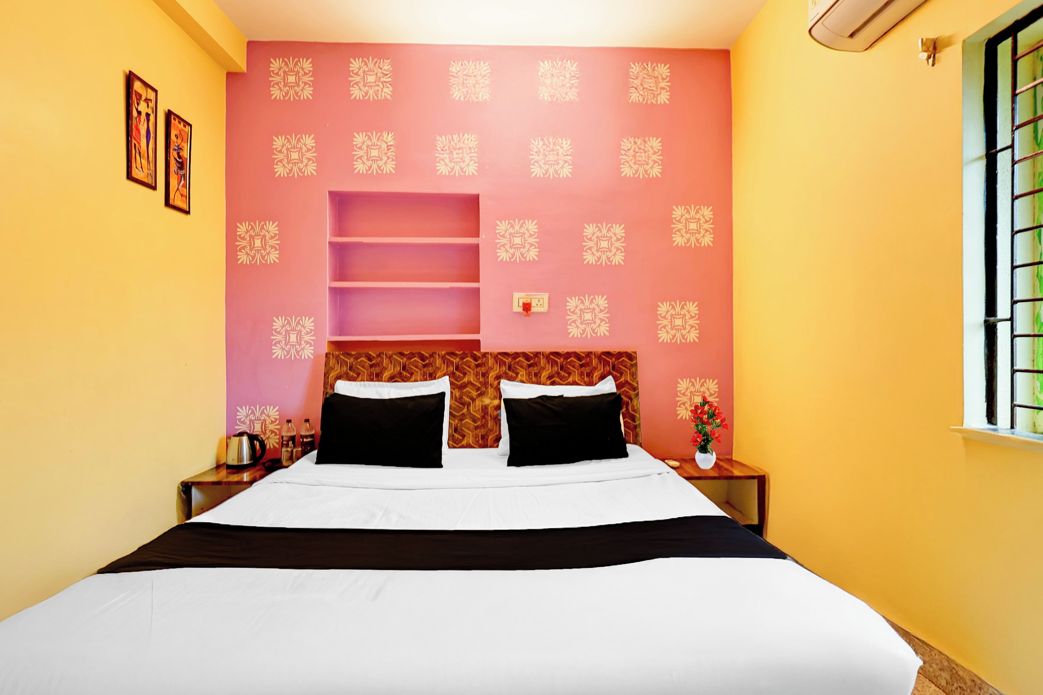 Habitación de hotel con cama, mesita de noche con hervidor, florero con flores y ventana con barrotes.