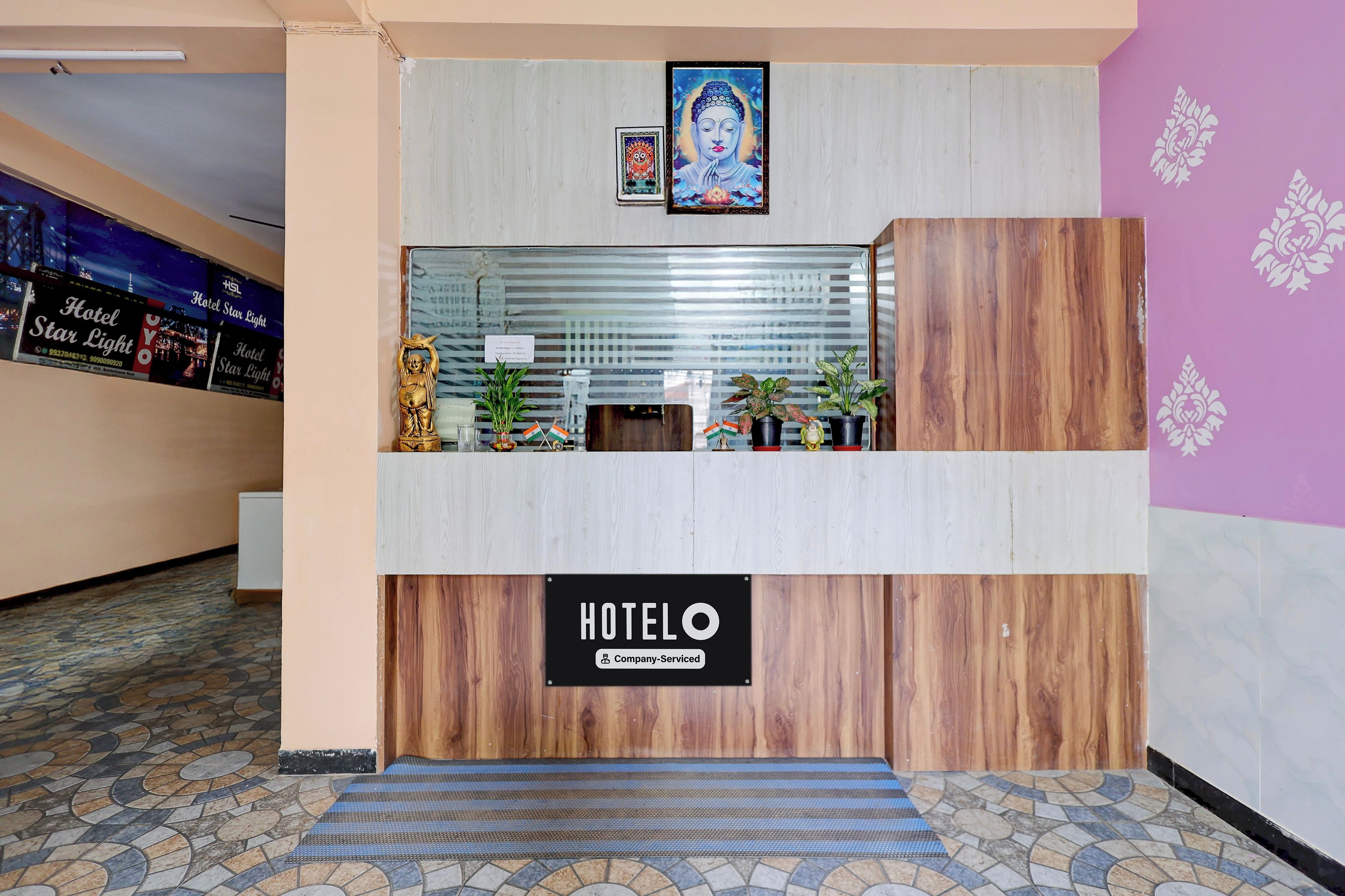 A hotel reception desk with a sign indicating it is company-serviced. There are potted plants and a small statue on the desk.