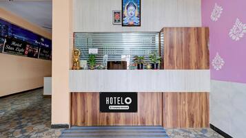 A hotel reception desk with a sign indicating it is company-serviced. There are potted plants and a small statue on the desk.