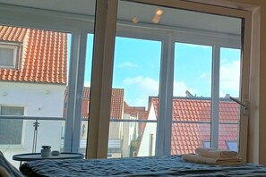 A room with a bed, a small table with a plant, and a view of neighboring buildings through large windows. - Holiday apartment Strandhaus (Norderney)