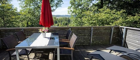 An outdoor deck with a table and chairs, a red umbrella, and a view of trees and hills.