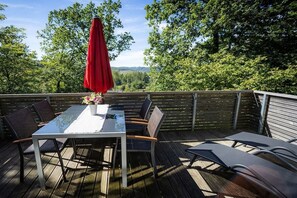 An outdoor deck with a table and chairs, a red umbrella, and a view of trees and hills. - Holiday home in Brilon with swimming pool (Brilon)