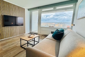 Ein modernes Wohnzimmer mit einem an einer Holzvertäfelung montierten Flachbildfernseher, einem Sofa mit einem türkisfarbenen Kissen, einem kleinen Holztisch und einem großen Fenster mit Blick auf Gebäude.