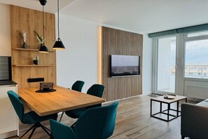 Ein modernes Wohnzimmer mit einem Holzesstisch, smaragdgrünen Stühlen, einem Flachbildfernseher und einem großen Fenster mit Blick auf die Stadt.
